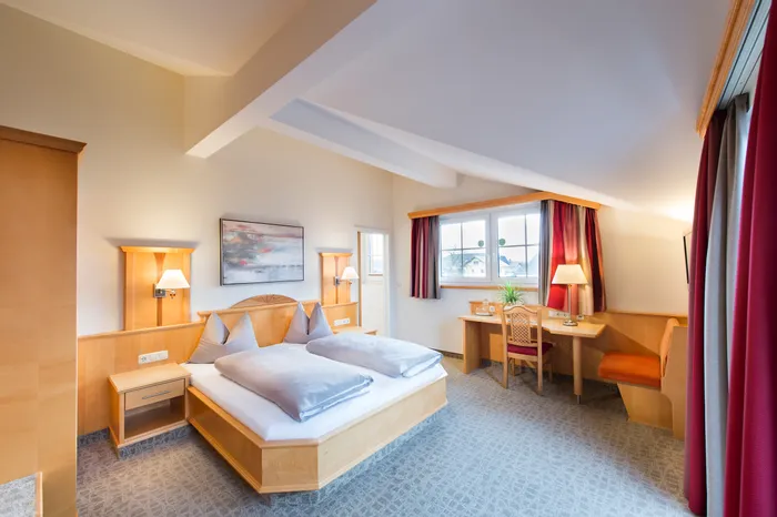 Bright hotel room with double bed, desk, and large window with red curtains